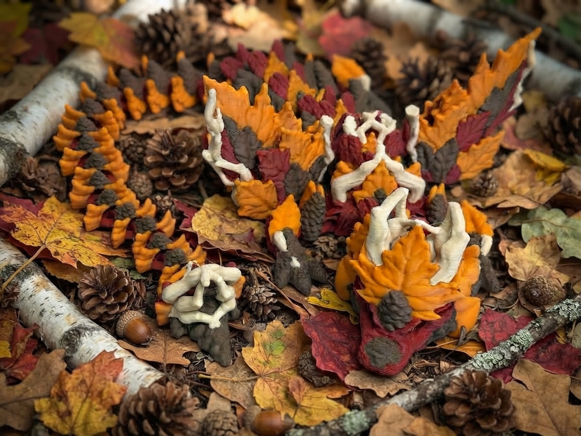 A 3D-printed articulated dragon figurine styled with autumn leaves and pinecones, featuring orange, red, brown, and cream colors. Displayed on a forest floor.